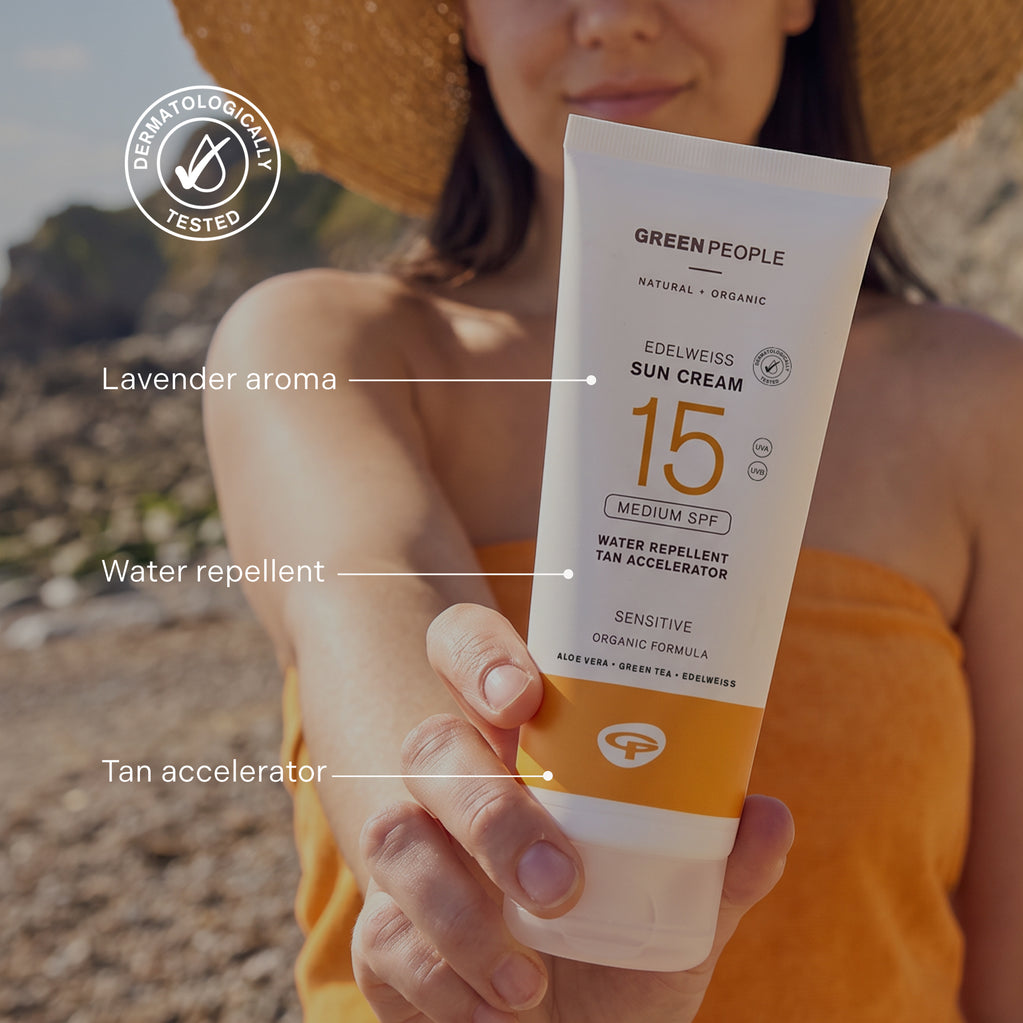 En hånd holder en tube Edelweiss Sun Cream med Tan Accelerator SPF15 på en solrik strand foran en kvinne med stråhatt.