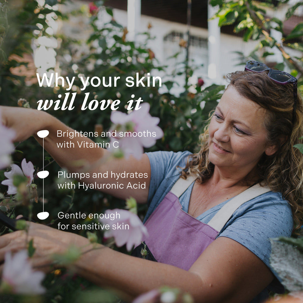 En smilende moden kvinne steller i hagen utendørs nær rosa blomster, noe som fremhever fordelene med Age Defy+ Collagen Boost Vitamin C Serum.